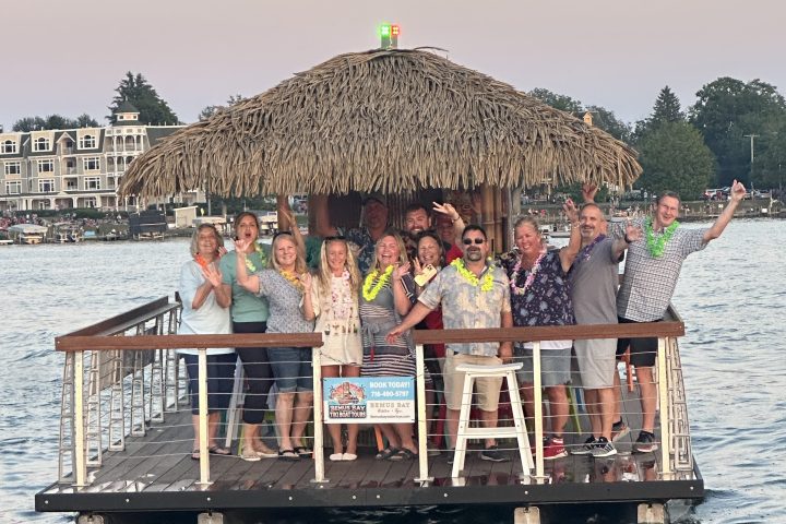 Group of people on a tiki bar boat on a lake with buildings in the background.
