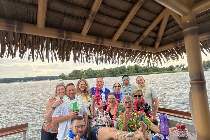 Group of people wearing leis, smiling and holding drinks under a tiki hut on a dock by the water.