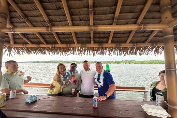 Group of people in leis under a tiki hut on a lake.