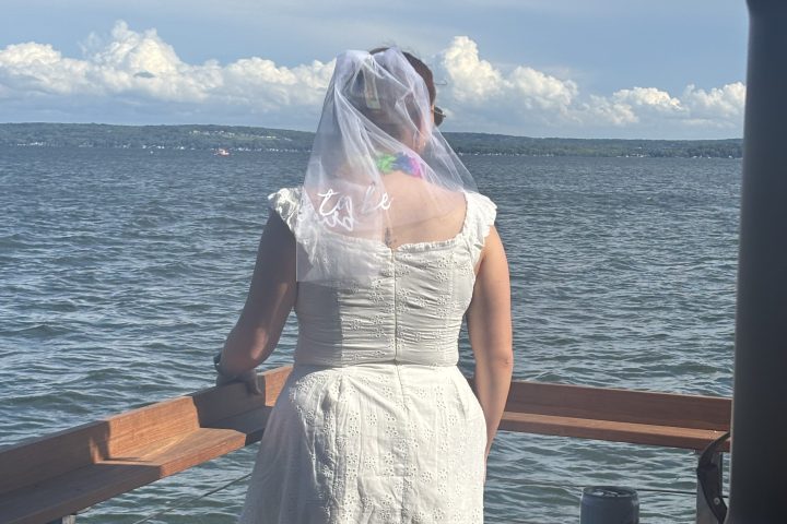 Woman in white dress with veil on dock gazing at lake under thatched roof.