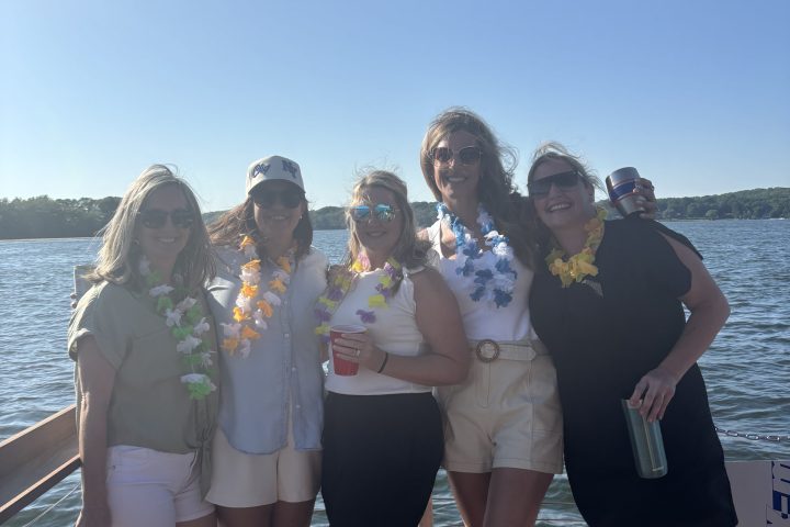 Five people in leis standing on a dock by the water under a straw canopy, smiling and enjoying a sunny day.