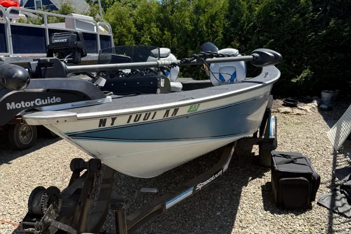 Boat on trailer in driveway next to trees, clear blue sky.