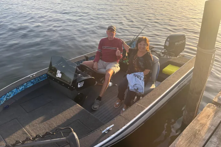 Two people sitting in a small boat on a calm lake near a dock.